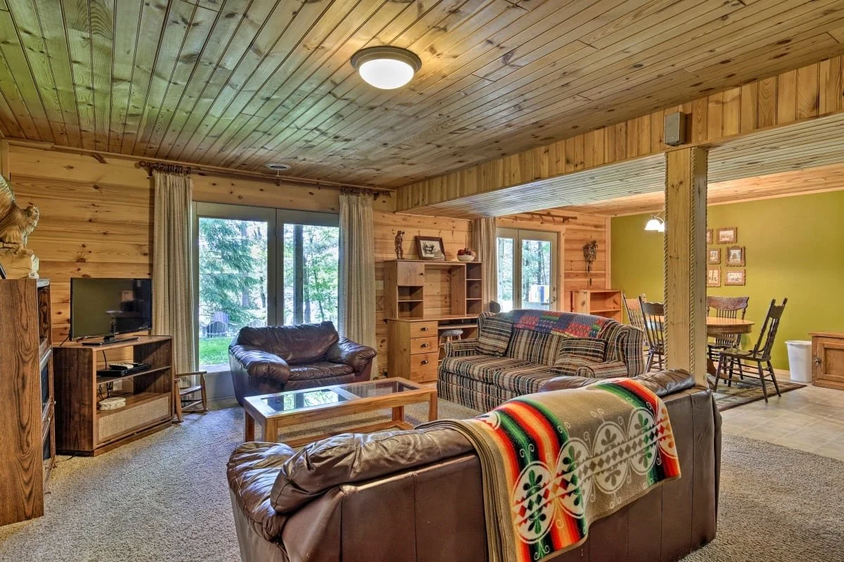 A living room with wood-paneled walls and ceiling, featuring a brown leather sofa, a plaid fabric sofa, a glass coffee table, and a wooden TV stand with a flat-screen TV. There are large windows with beige curtains letting in natural light, and a din