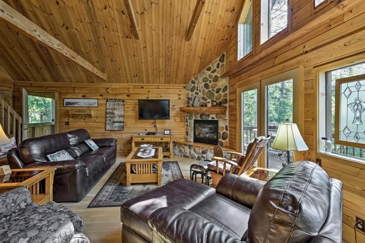 Living room with wooden walls and ceiling, stone fireplace, leather sofas, a patterned armchair, a coffee table, and large windows showing a wooded outdoor area.