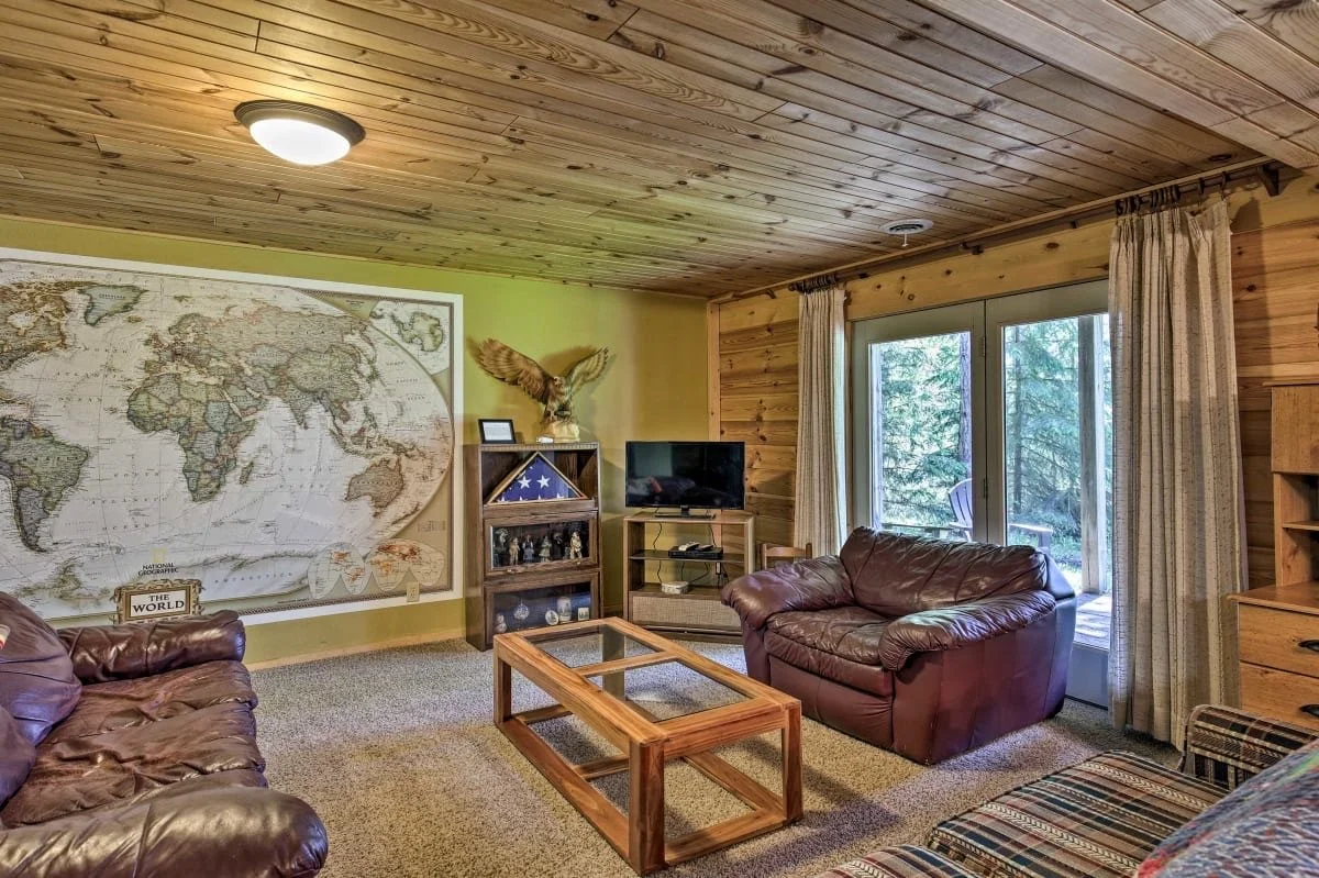 Living room with leather sofas, a large world map on the wall, a wooden coffee table, TV on a stand, a wooden bookshelf, and sliding glass doors leading outside to trees.