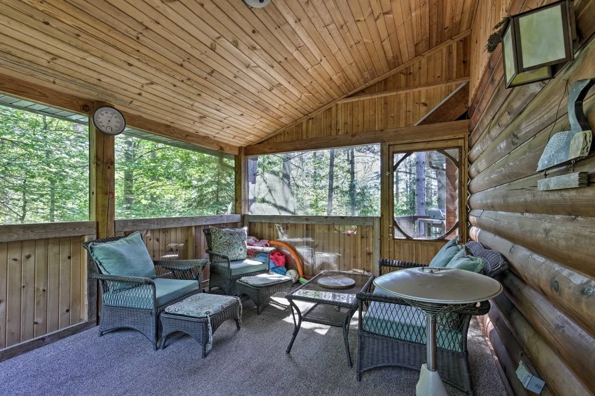 Screened porch with wicker furniture, including armchairs, a small table, and a larger coffee table, surrounded by wooded trees visible through the screened windows.