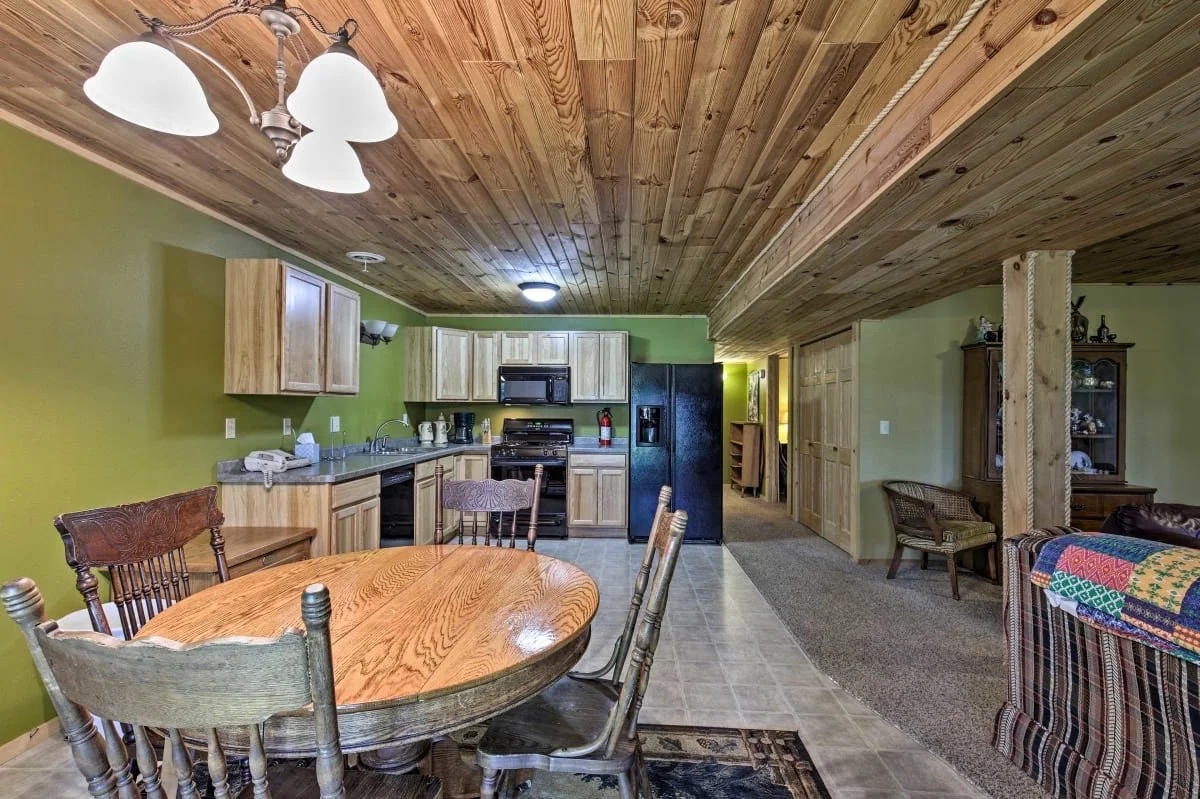 Interior of a rustic kitchen and dining area with wooden ceiling and cabinetry, green walls, a round wooden table with chairs, black refrigerator, stove, microwave, and various kitchen appliances. In the background, a hallway with carpeted floor and 