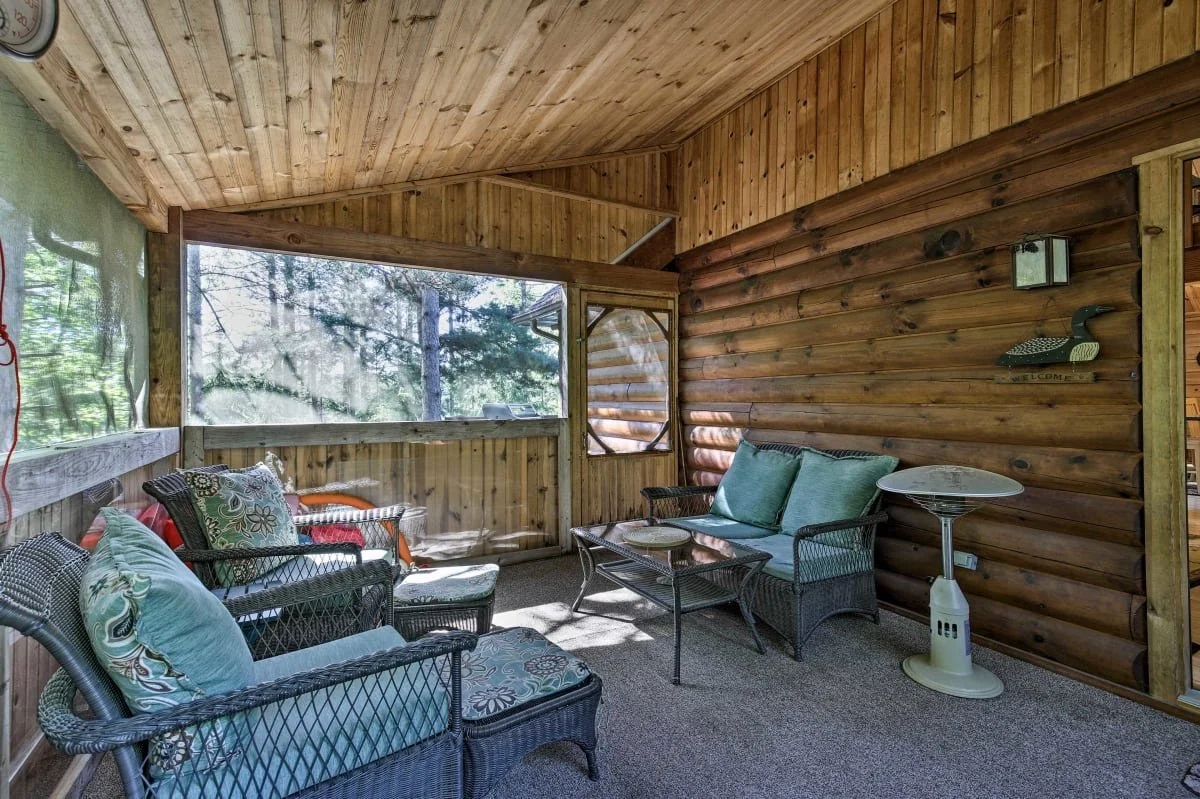 Covered porch area with wicker furniture, cushions, and a small side table, surrounded by wooden walls and large windows showing trees outside.