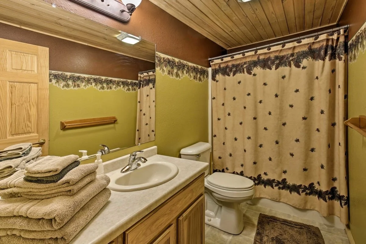 Bathroom with a wooden ceiling, green and brown walls, a large mirror, a sink with a countertop, a stack of towels, a toilet, a shower with a curtain featuring pinecones and pine branches, and a small brown rug.