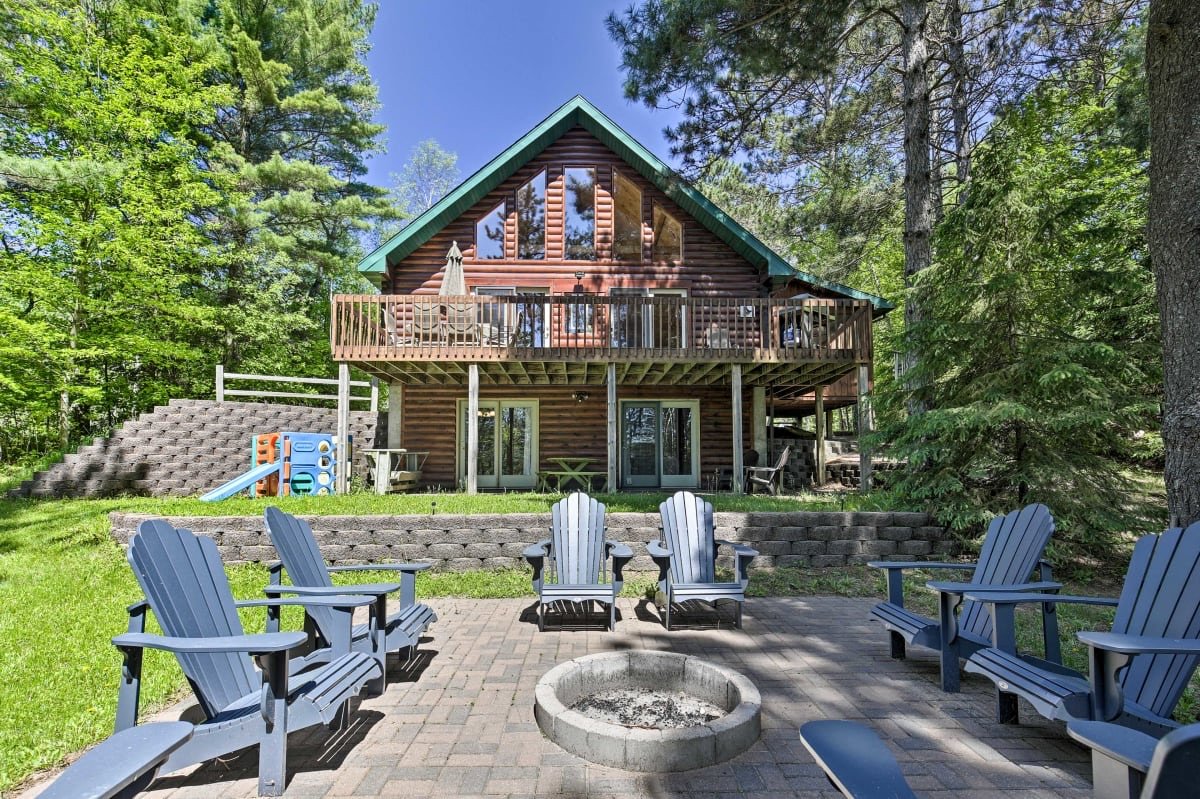 A wooden cabin with a high-pitched roof, large front windows, and a wrap-around balcony in a forested area. The backyard has outdoor chairs surrounding a fire pit on a paved patio, and there are trees and a small playset with slides nearby.