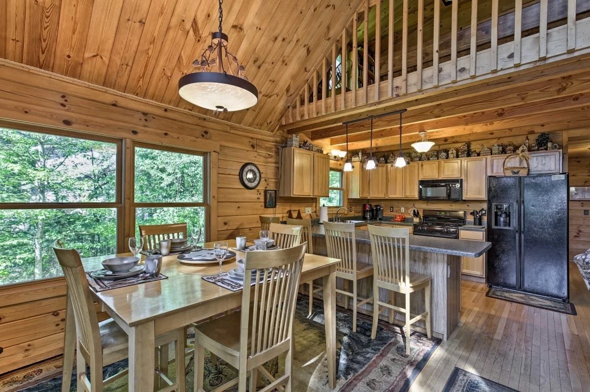 Wood-paneled kitchen and dining area with large window overlooking trees. Dining table set with dishes and glasses. Kitchen with bar stools, cabinets, black refrigerator, microwave, stove, and pendant lighting.