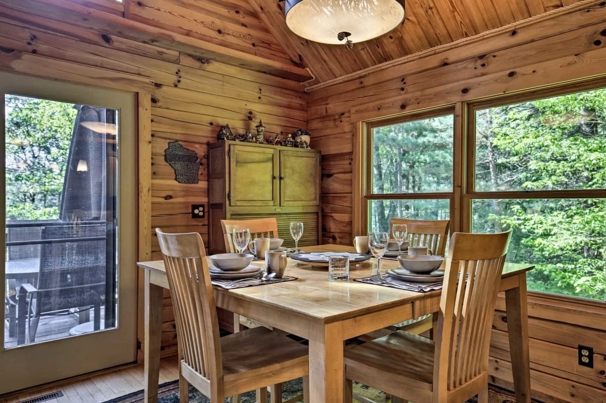 A dining area with a wooden table set with plates, glasses, and cups, inside a room with wood-paneled walls and large windows showing green trees outside.