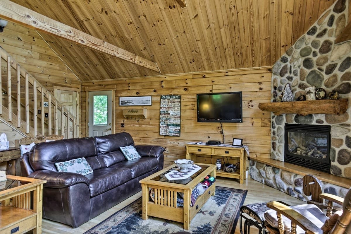 Interior of a cozy living room with wooden walls and a vaulted ceiling, featuring a brown leather sofa, a wooden coffee table, a stone fireplace with a wooden mantle, and a wall-mounted flat-screen TV.