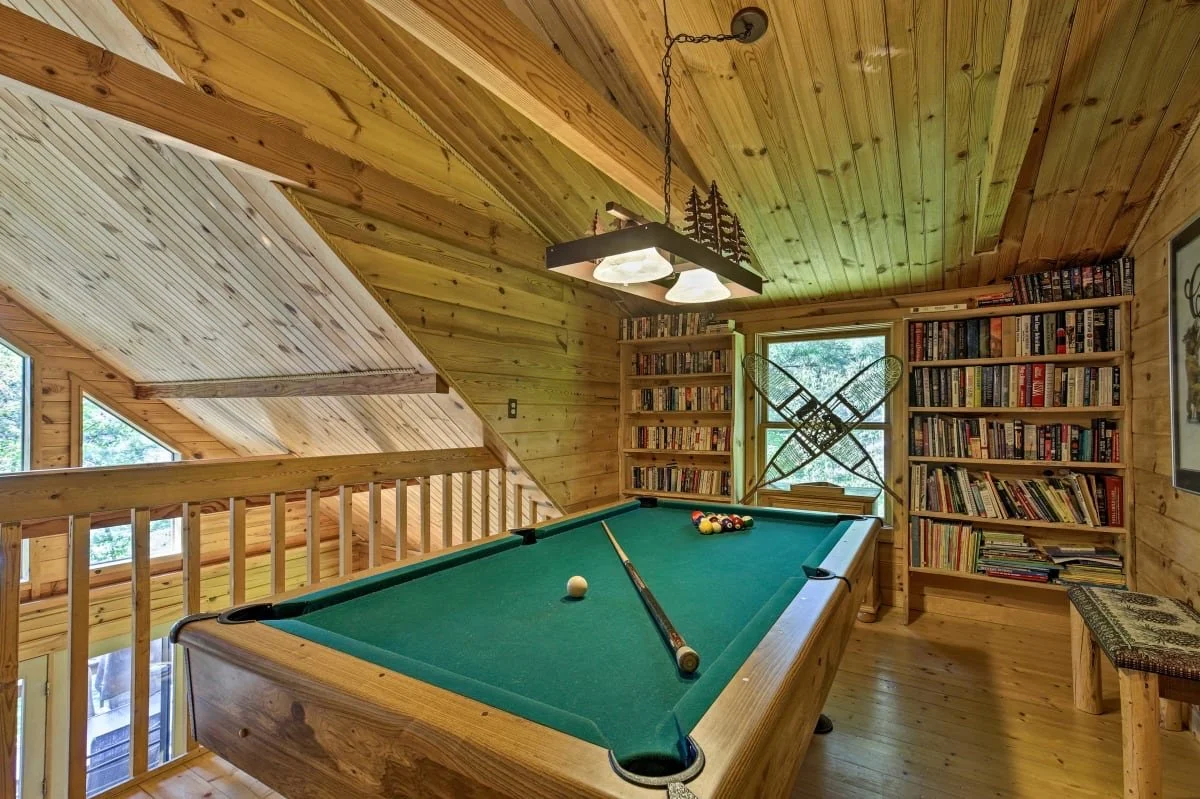 A rustic indoor game room with a pool table, bookshelves filled with books, snowshoes hanging on the wall, and a window showing greenery outside. The room features wooden walls, ceiling, and flooring, with a chandelier overhead.