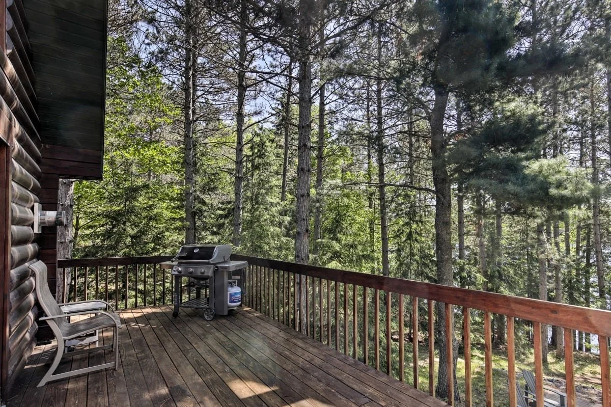 Wooden balcony with two chairs, a gas grill, and a forest of tall trees in the background.
