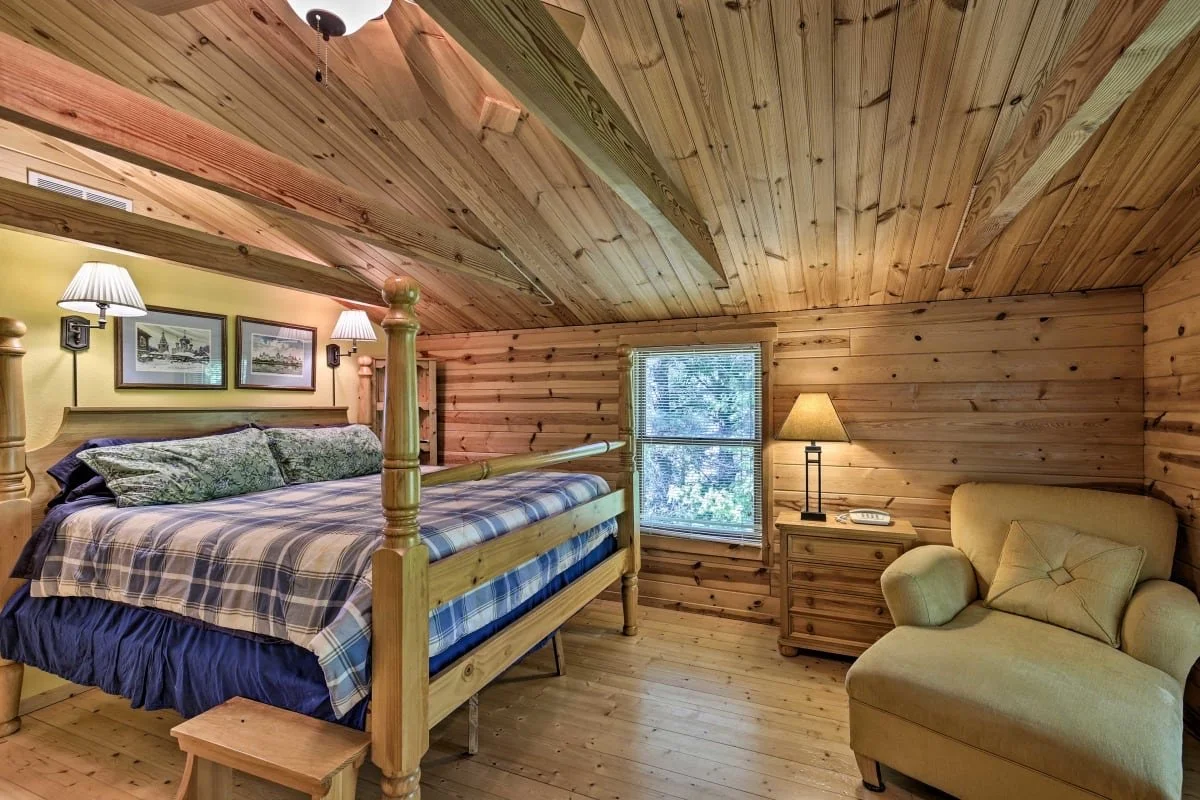 Cozy bedroom with wooden walls, ceiling, and floor. Features a large four-poster bed with plaid bedding and green decorative pillows, two wall sconces, and framed pictures above the headboard. A window with blinds and a nightstand with a lamp and pho