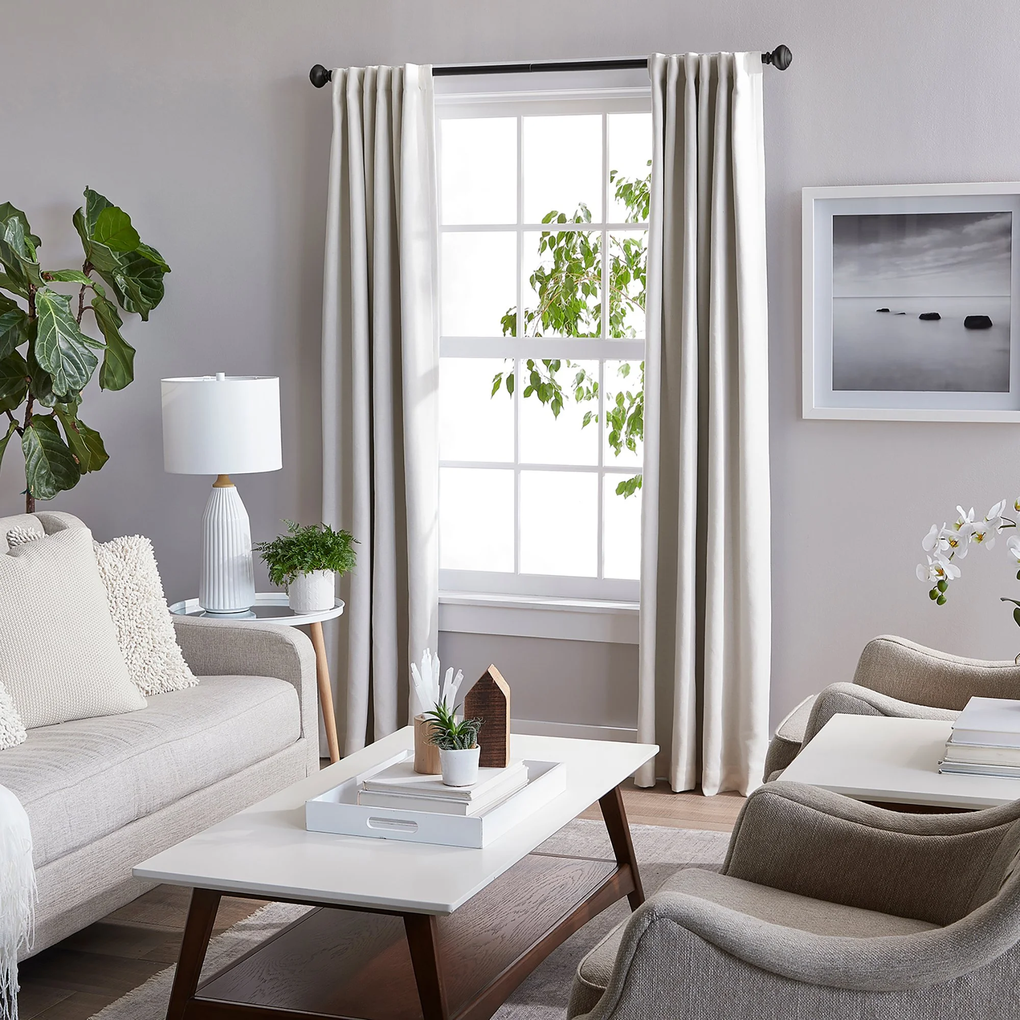 Living room with white curtains, beige sofa, armchair, coffee table with decorative items, large window with black curtain rod and white drapery, framed landscape photo, green plant, and side table with lamp.