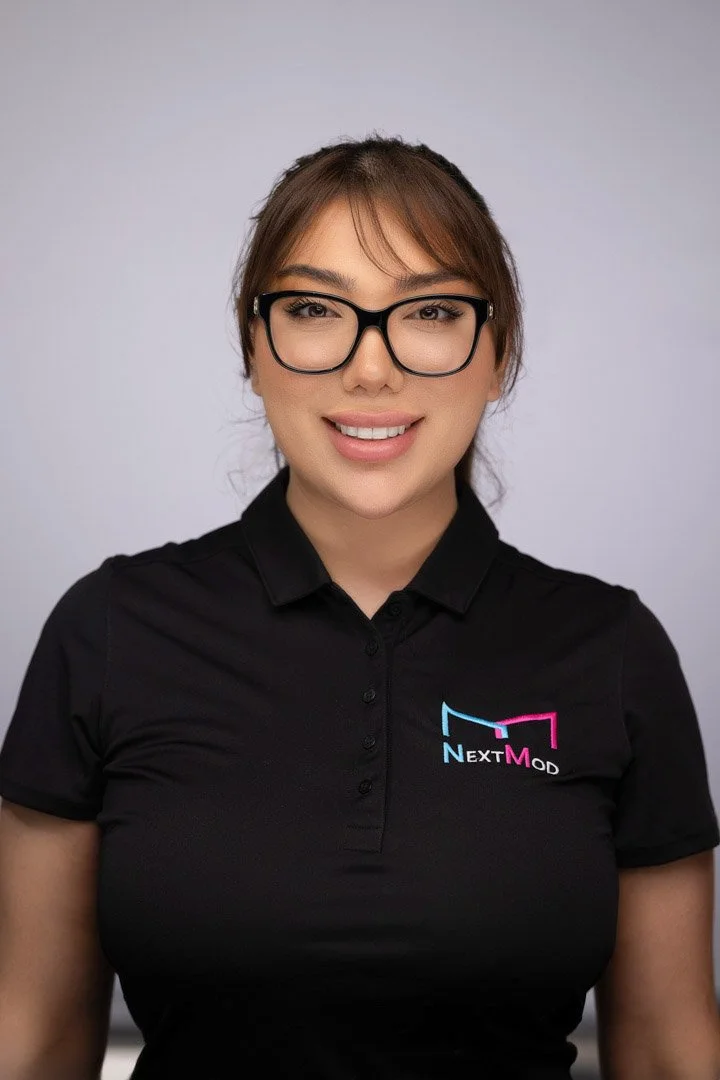 A woman smiling, wearing glasses and a black polo shirt with a logo that says 'NextMod' on the left side of her chest.