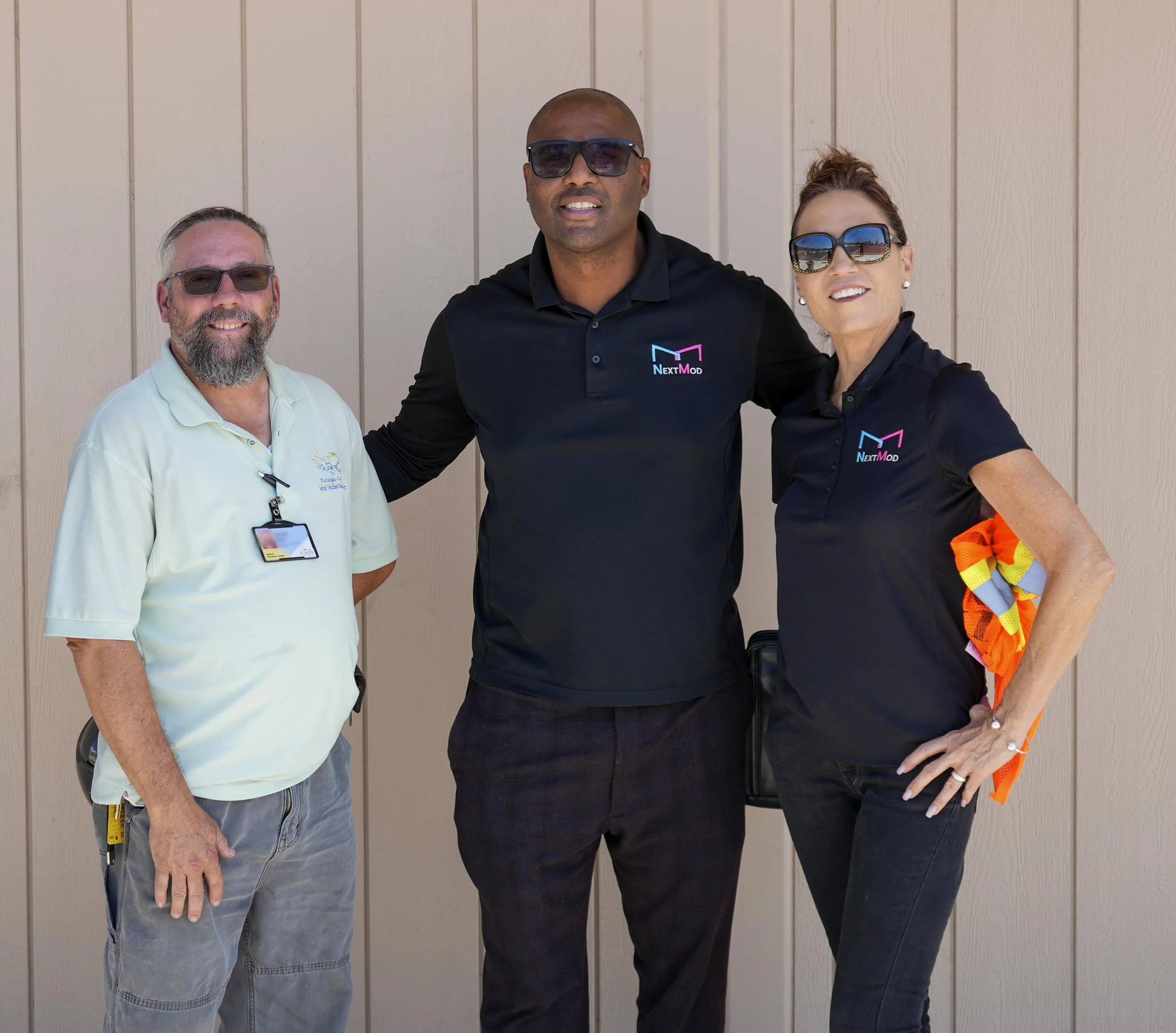 Three people standing together outdoors in front of a beige wall, smiling, wearing sunglasses, with two of them wearing black company shirts with a logo.