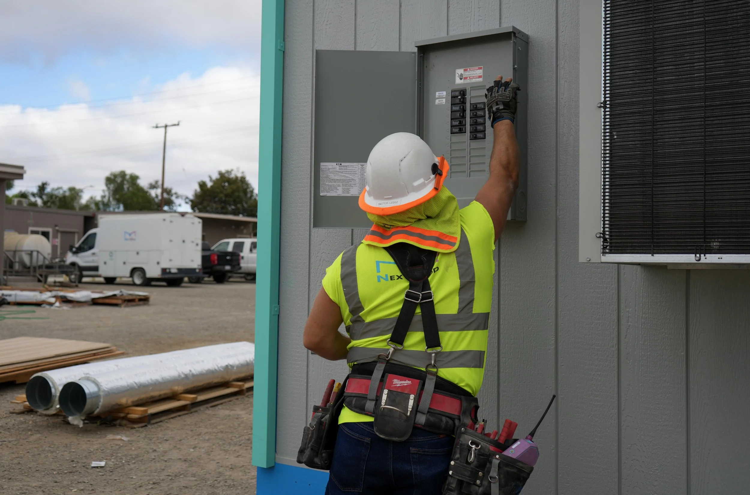 A construction worker wearing a white safety helmet, neon yellow safety vest, and gloves working on an electrical panel outside a building.