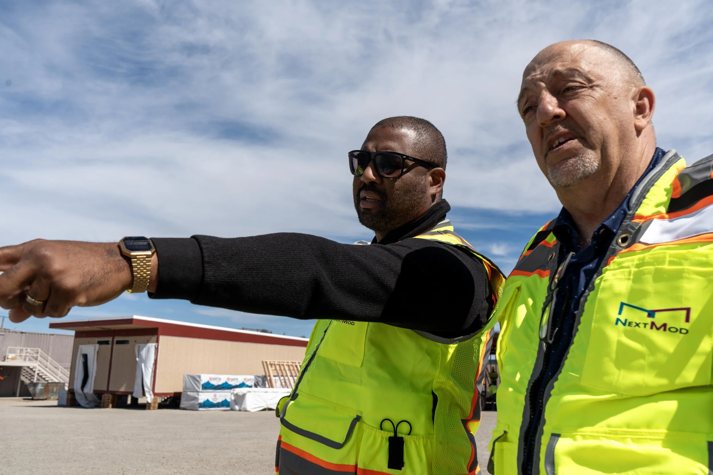 Two men in bright yellow safety vests standing outdoors on a sunny day with a building and some equipment in the background. One man is pointing while the other looks on.
