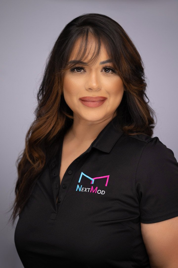 A woman with long wavy brown hair and a warm smile wearing a black polo shirt with a 'Next Mod' logo.