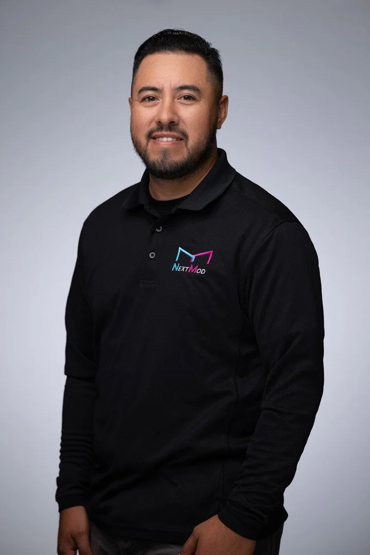 A man wearing a black polo shirt with a 'NextMod' logo, standing in front of a light gray background, smiling at the camera.