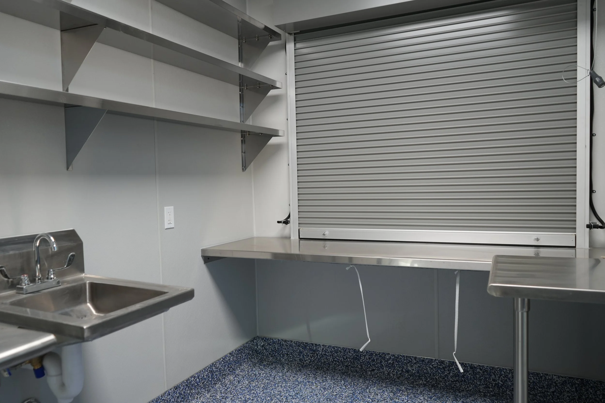 Empty stainless steel workspace with shelves, a sink, and a closed roll-up window in a commercial kitchen or food prep area.