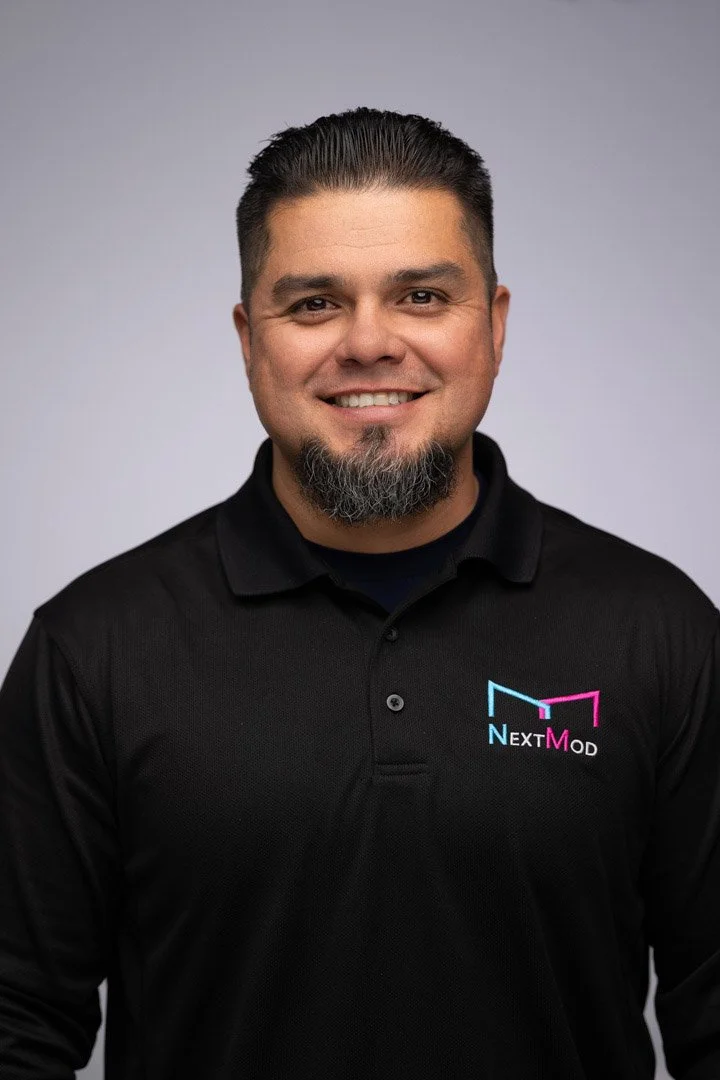 A man with short dark hair and a goatee, wearing a black collared shirt with a 'NextMod' logo, smiling at the camera against a plain gray background.