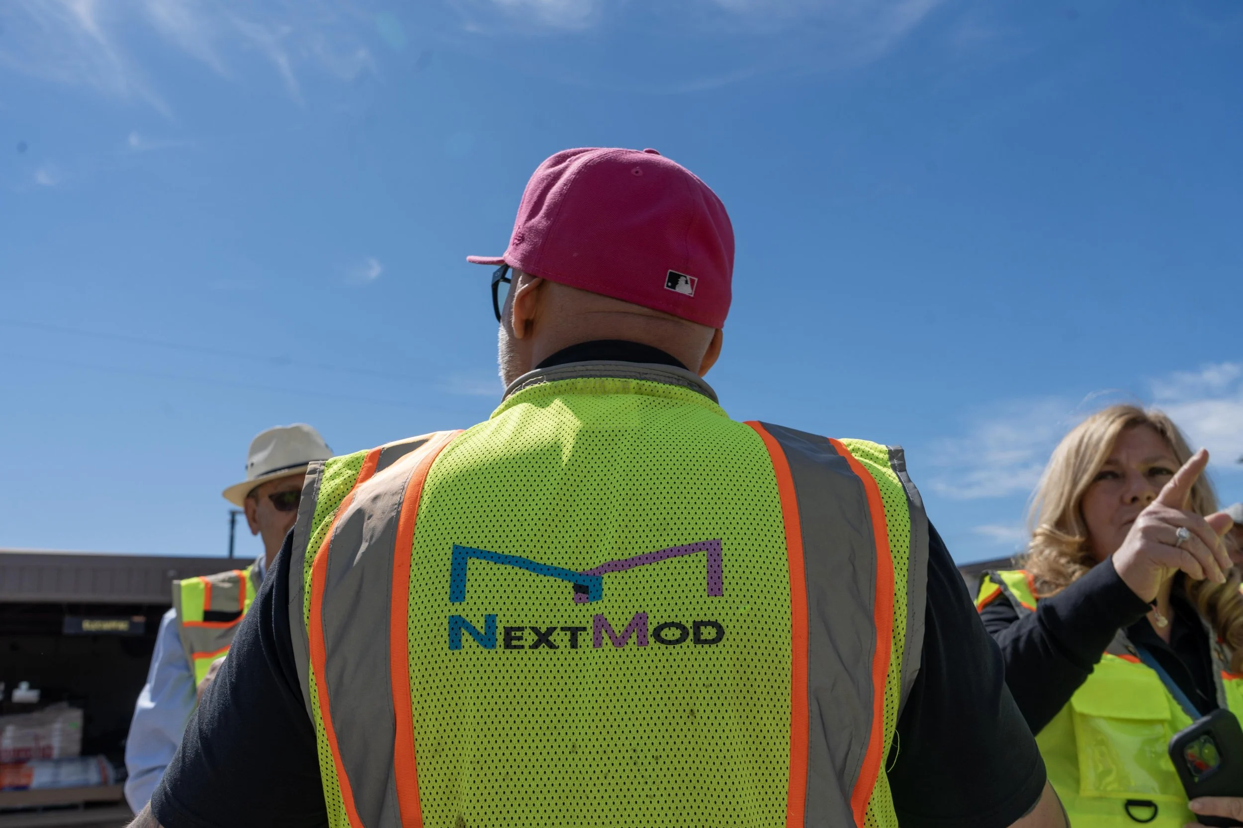 A group of people wearing yellow safety vests and hats outdoors under a clear blue sky. The person in the foreground has their back to the camera, with the logo 'Next Mod' on their vest, and is wearing a pink cap and sunglasses.