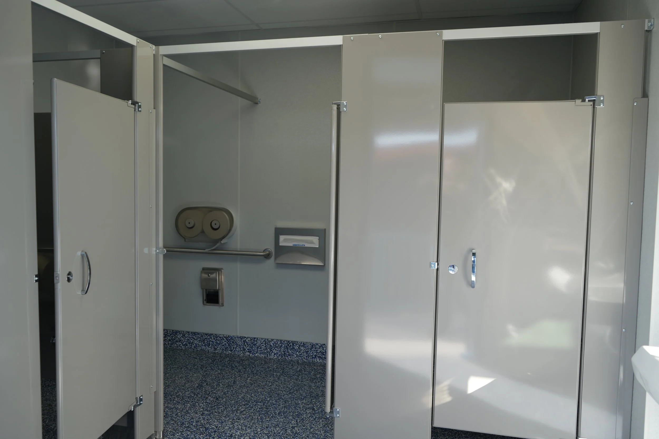 Empty unisex accessible restroom stall with open doors, equipped with handrails, paper towel dispenser, and trash bin, with gray walls and blue speckled flooring.