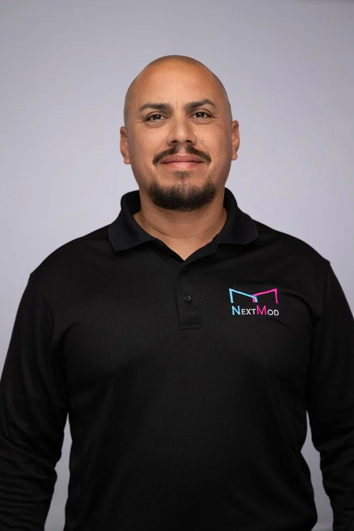 A man with a shaved head and goatee wearing a black polo shirt with a 'NextMod' logo, standing against a plain gray background.