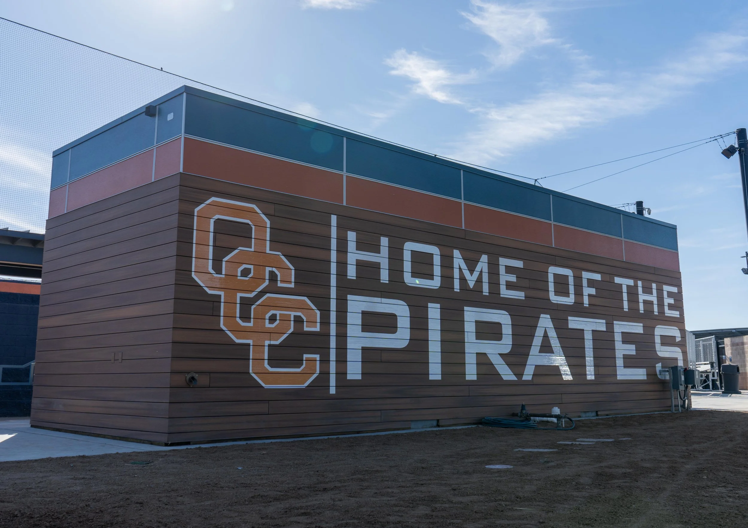 A building with a wall featuring a large sign that reads 'HOME OF THE PIRATES' with a skull and crossbones logo, under a partly cloudy blue sky.