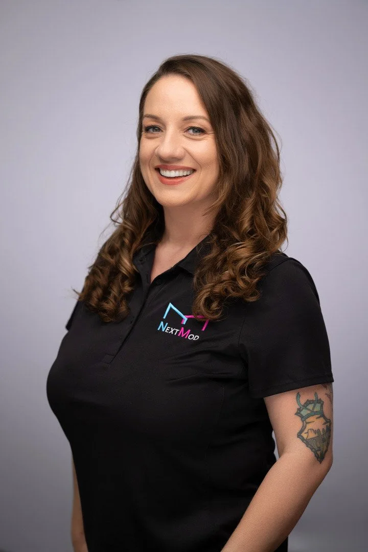 A woman with long, curly brown hair, smiling, wearing a black polo shirt with the NextMod logo, and a tattoo on her left arm, standing against a plain gray background.