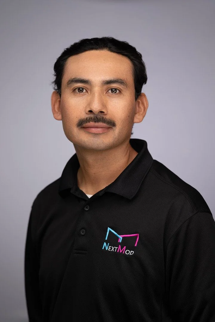 A man with a mustache and dark hair, wearing a black collared shirt with a 'Next Mod' logo, posing against a light gray background.