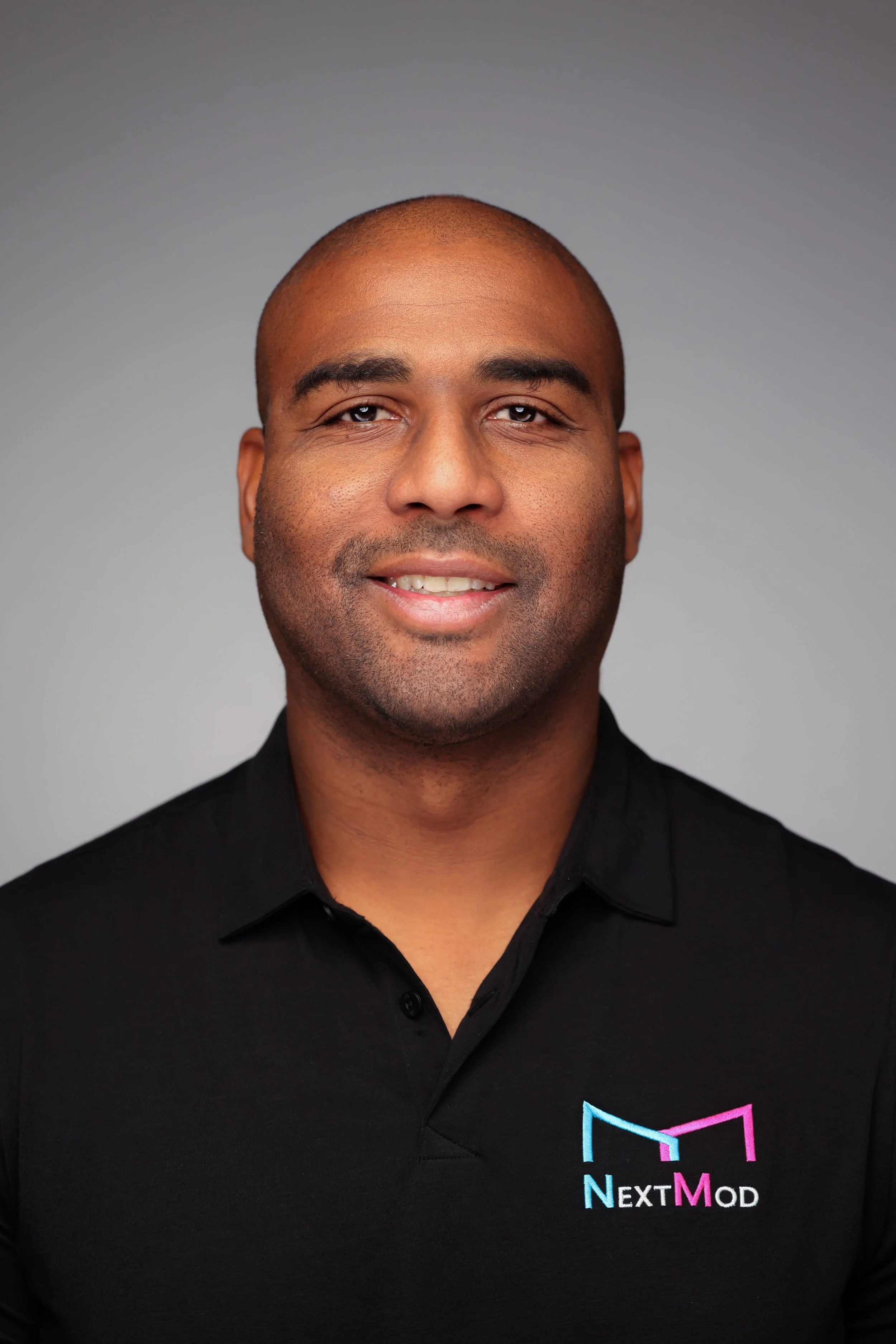 Headshot of a man with dark skin, wearing a black polo shirt with a logo that reads 'NextMod' in blue, pink, and white, against a gray background.