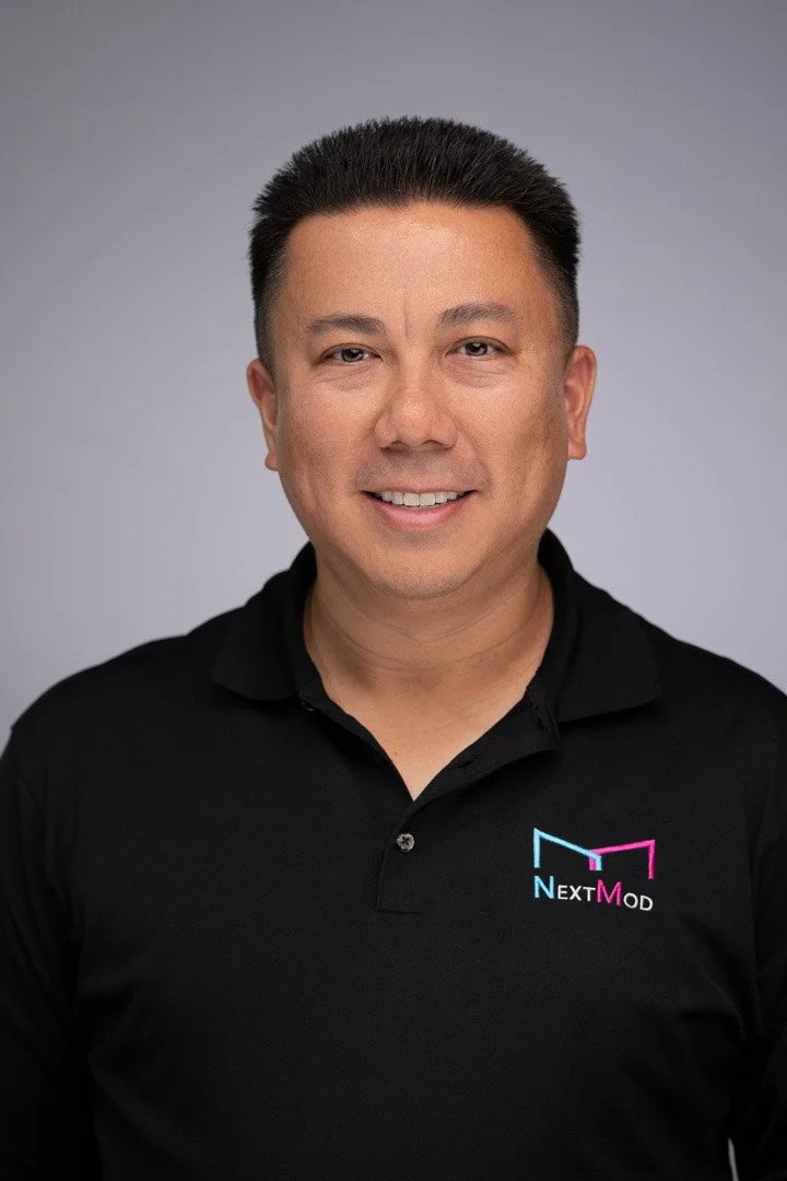 A man with short dark hair, wearing a black polo shirt with the NextMod logo, smiling at the camera against a plain gray background.
