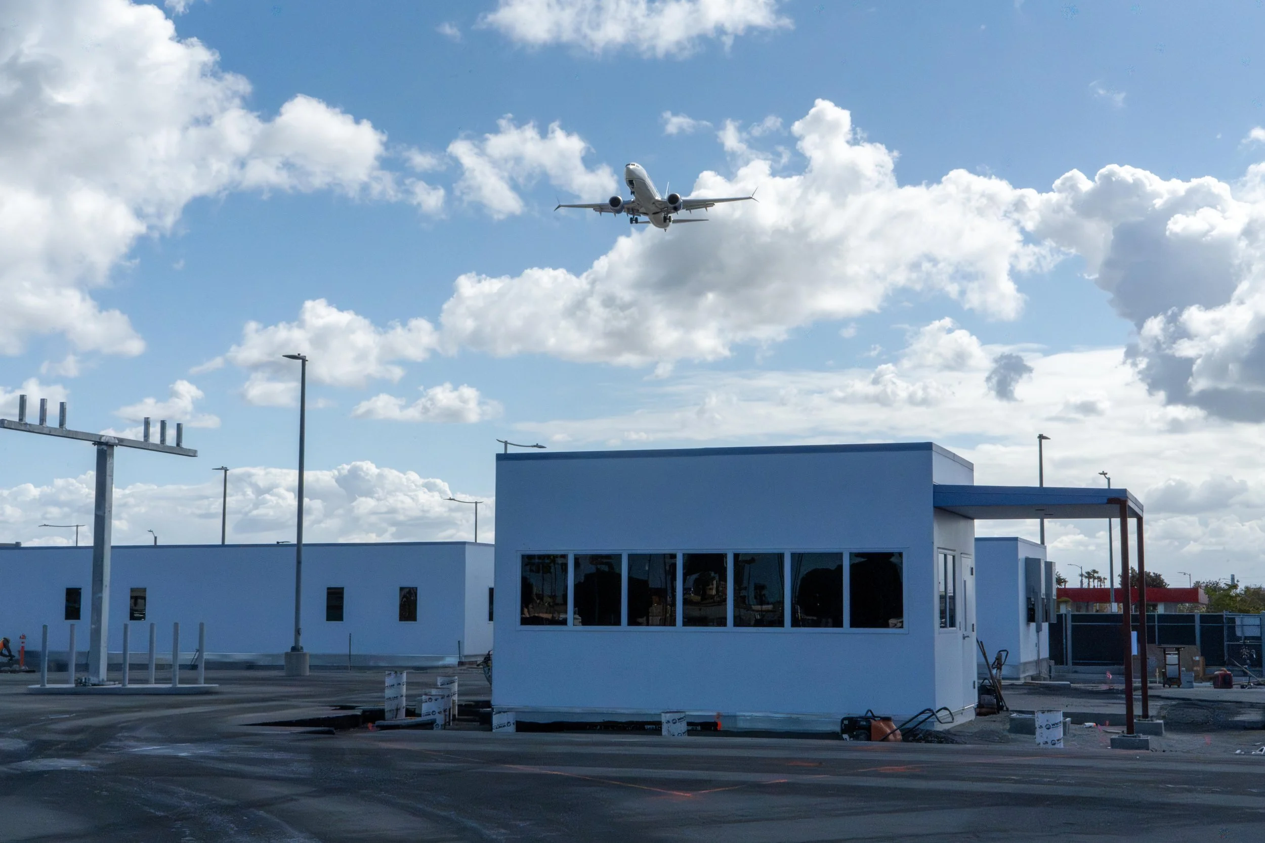 An airplane flying in the sky above a building with a white exterior, blue trim, and large windows, with clouds scattered across a blue sky.