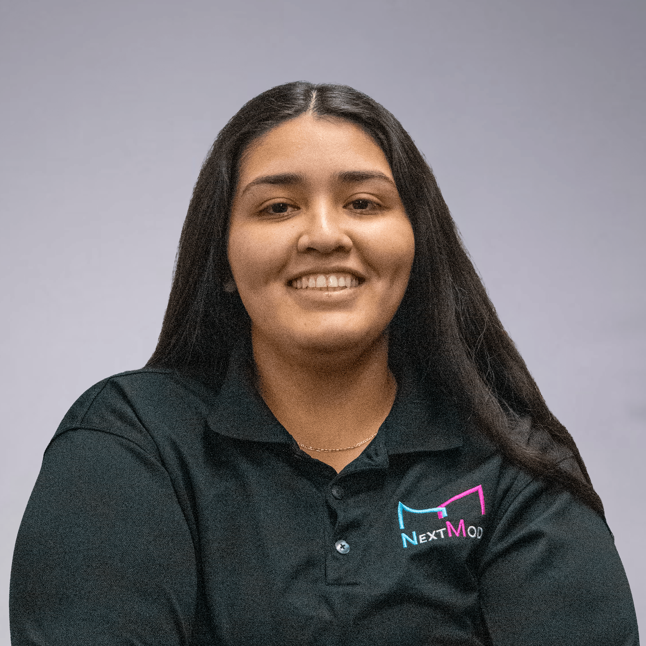 Young woman smiling, wearing a black polo shirt with a colorful 'NextMod' logo, standing against a plain gray background.