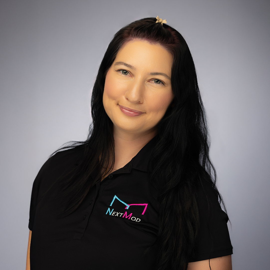 A woman with long wavy brown hair tied in two ponytails, wearing a black polo shirt with a logo that says 'NextMod', smiling at the camera against a plain gray background.