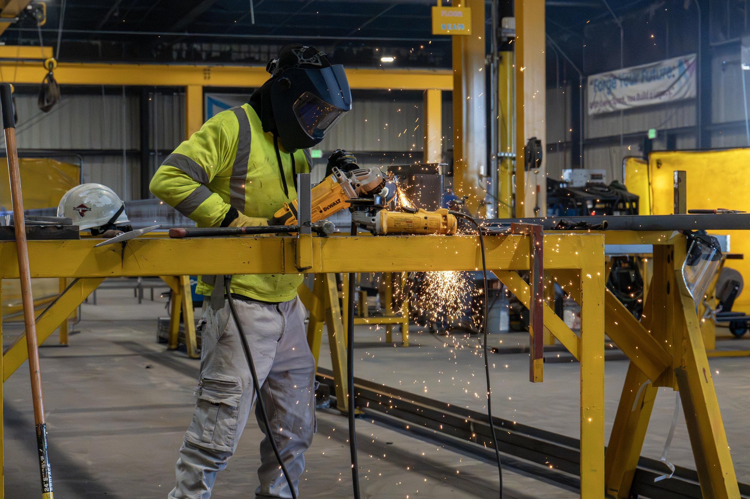 A worker wearing a yellow safety jacket, white pants, and a face shield welding metal inside a large industrial space with yellow equipment and scaffolding, sparks flying from the welding process.