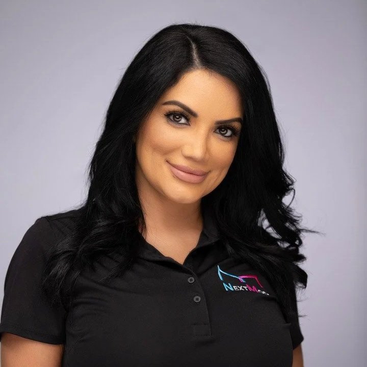 A woman with long black hair and makeup, wearing a black collared shirt with a colorful logo, smiling at the camera against a plain gray background.