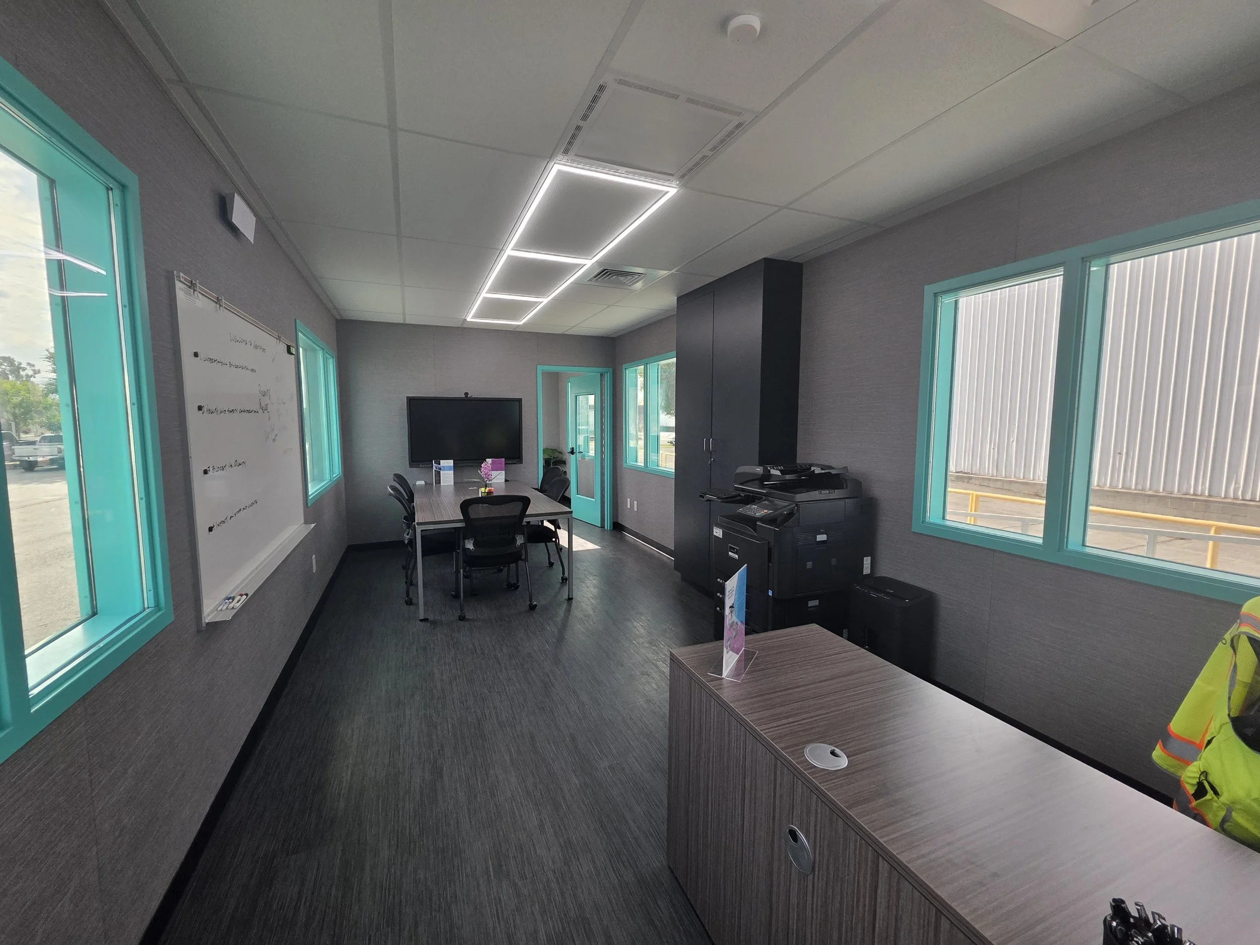Interior of the NextMod EcoSecure with a table, chairs, windows with teal trim, a whiteboard, and a large TV monitor on the wall.