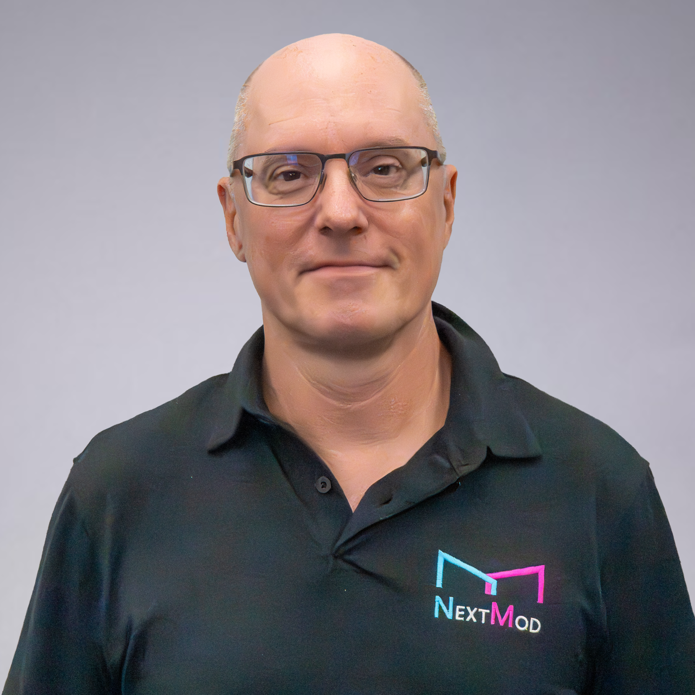 Headshot of a man with glasses, wearing a black polo shirt with a 'Next Mod' logo, standing against a plain light gray background.