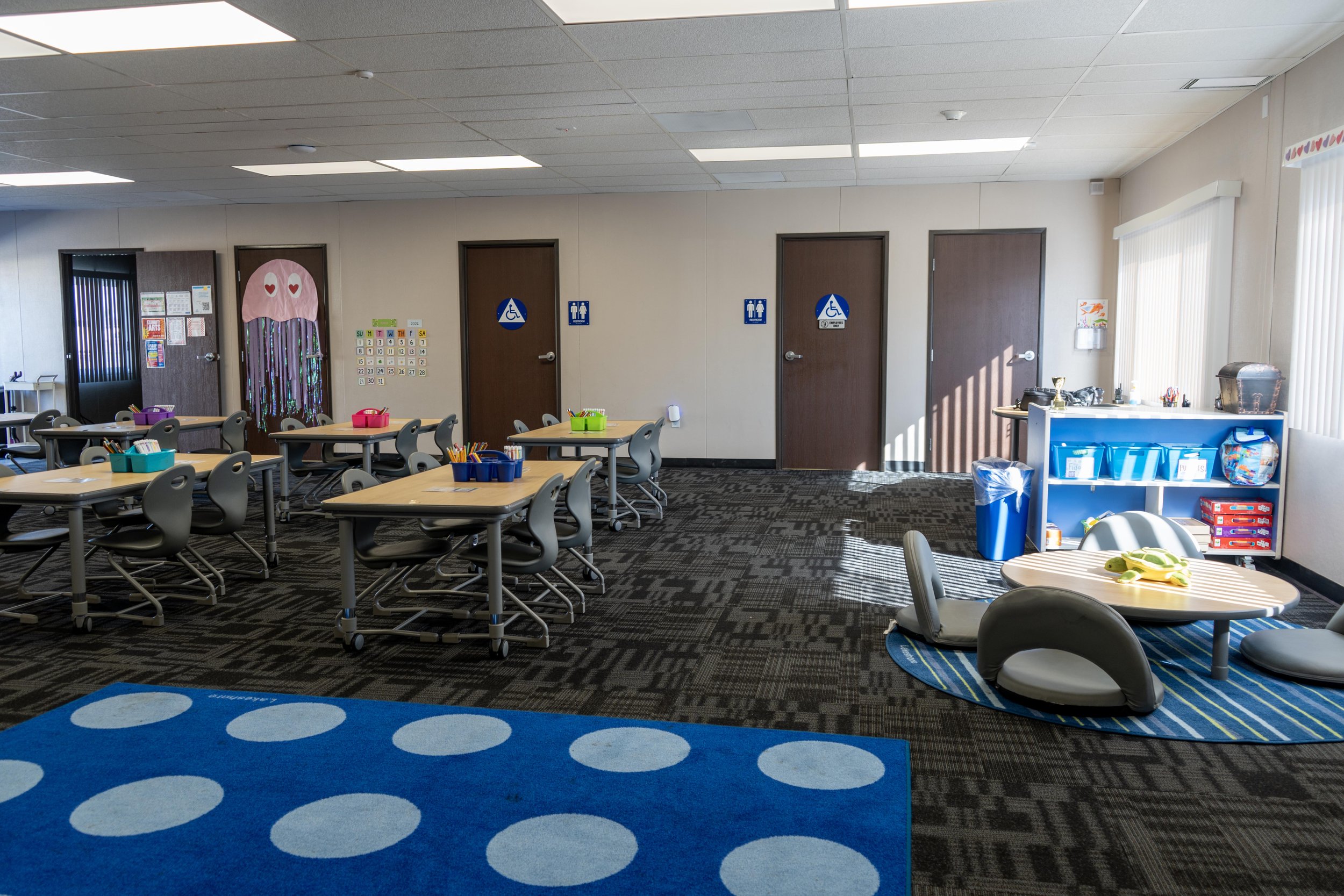 An empty classroom or activity room with tables and chairs, colorful bins with supplies, a blue rug with circles, a low table with cushions, and signs indicating accessible restrooms.