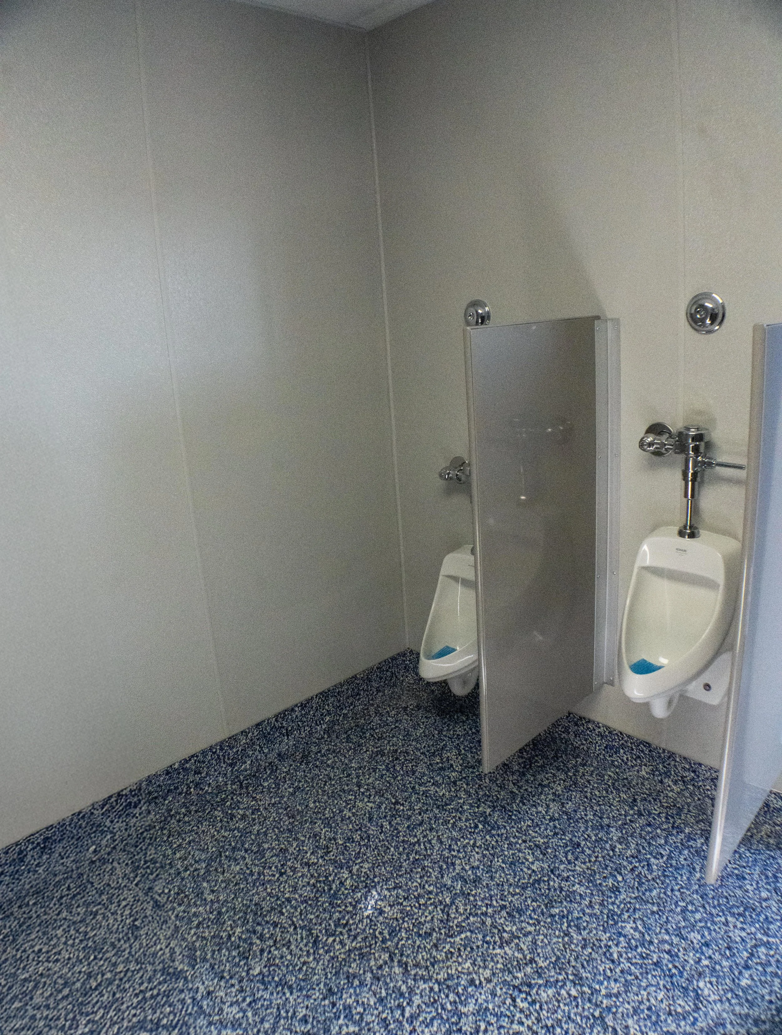 Public men's restroom with two urinals, one with a partition, on a speckled blue carpeted floor, plain gray walls, and no other fixtures visible.