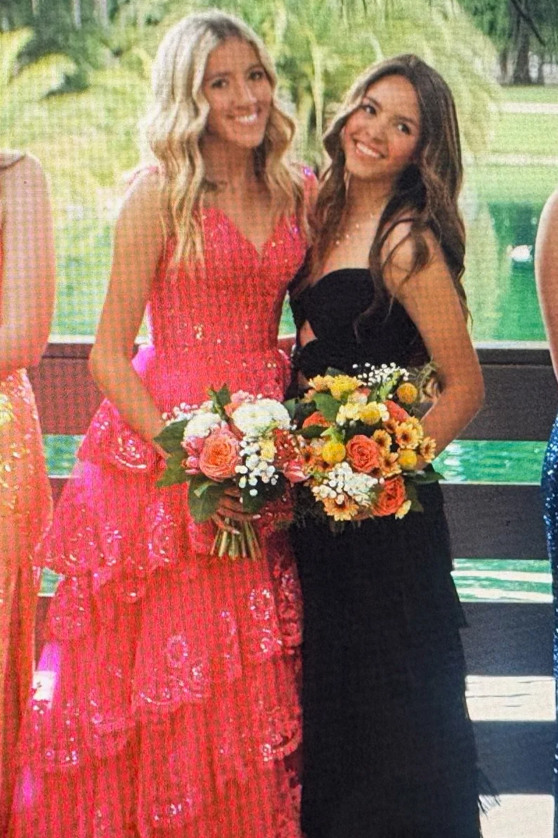 Two women in formal dresses holding bouquets, standing outdoors with a lake and trees in the background.