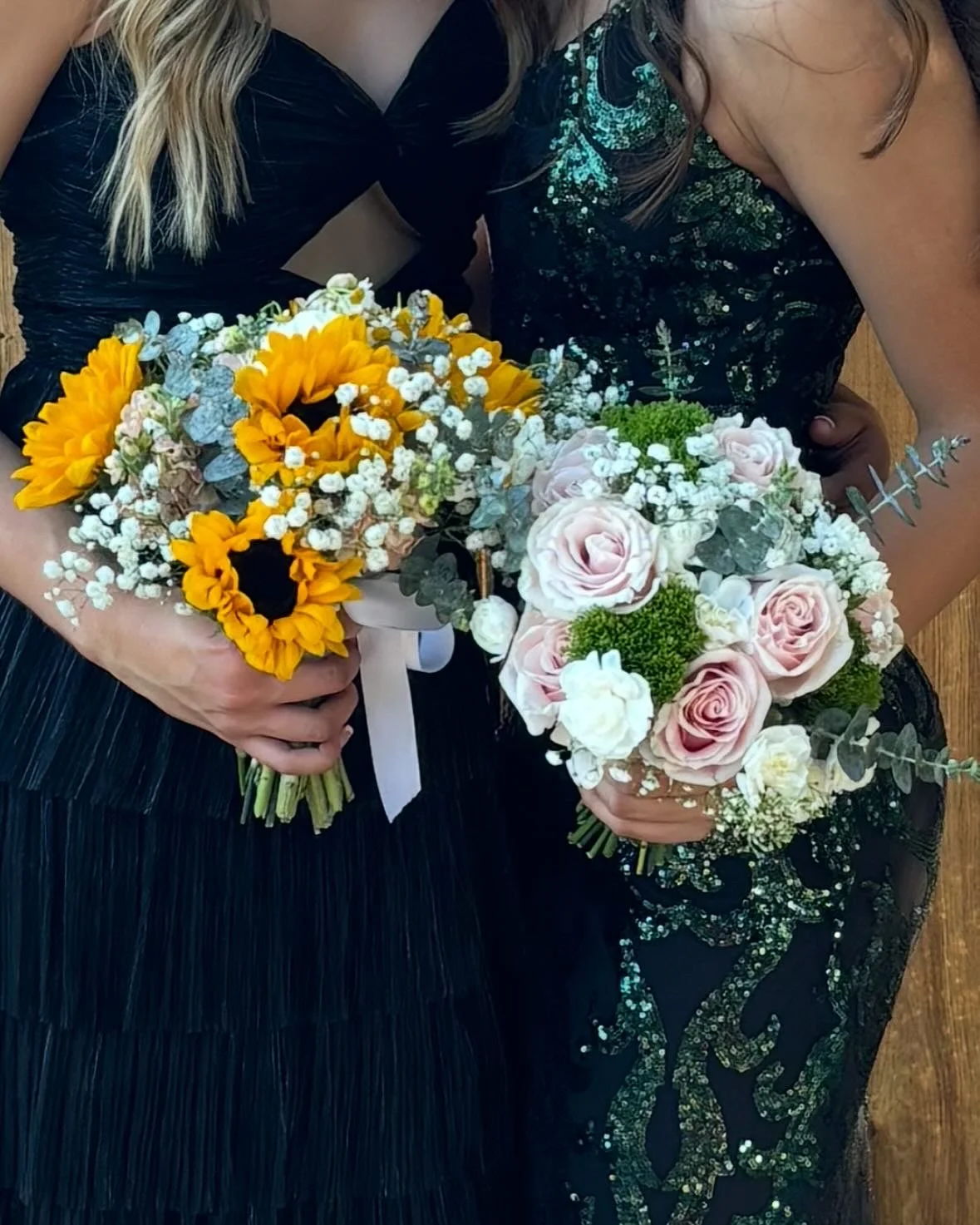 Two women holding bouquets of flowers, one with sunflowers and baby's breath, the other with pink roses, white flowers, greenery, and eucalyptus, in black dresses.