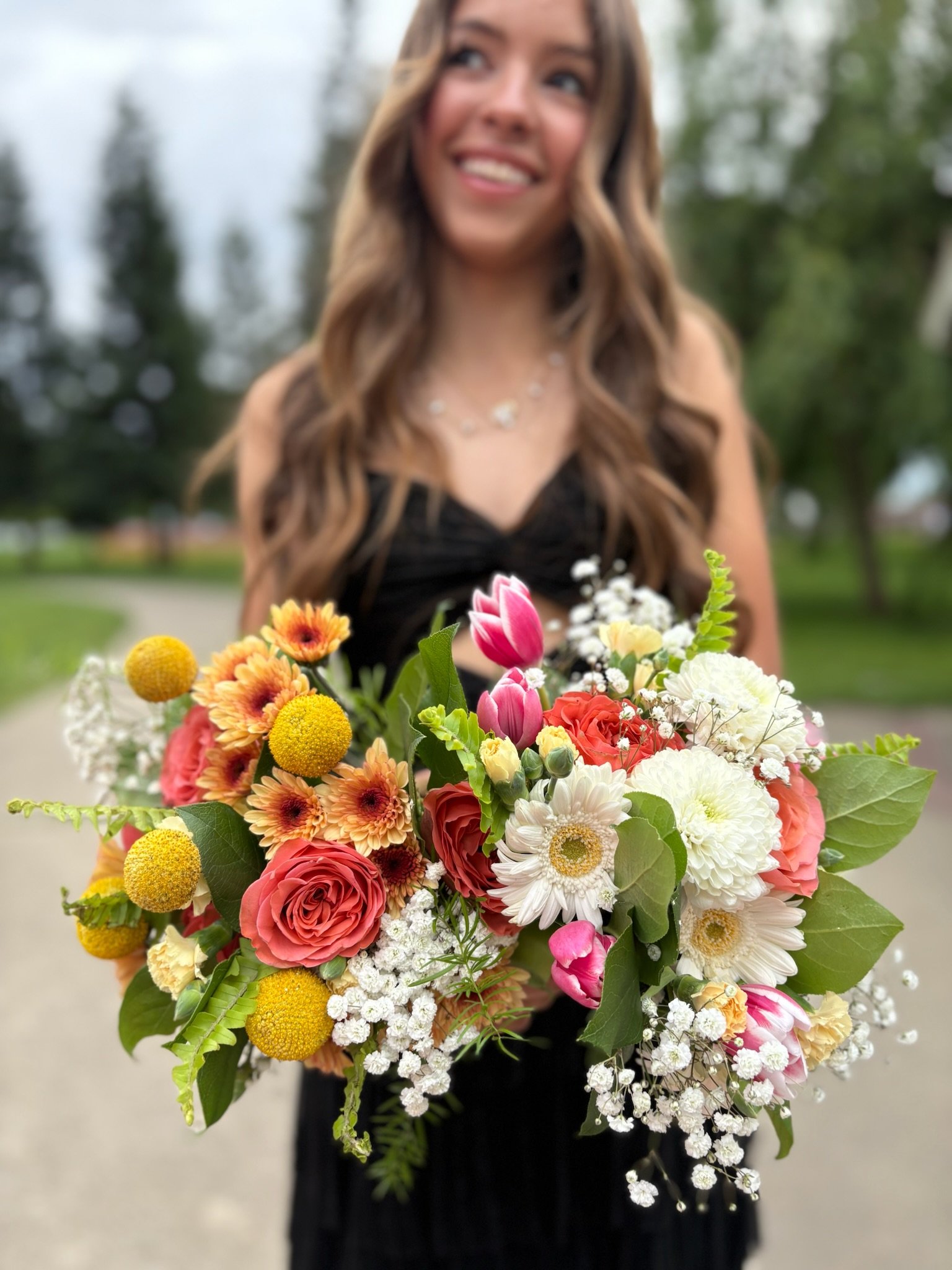A woman in a black dress holding a colorful bouquet of flowers outdoors, smiling.