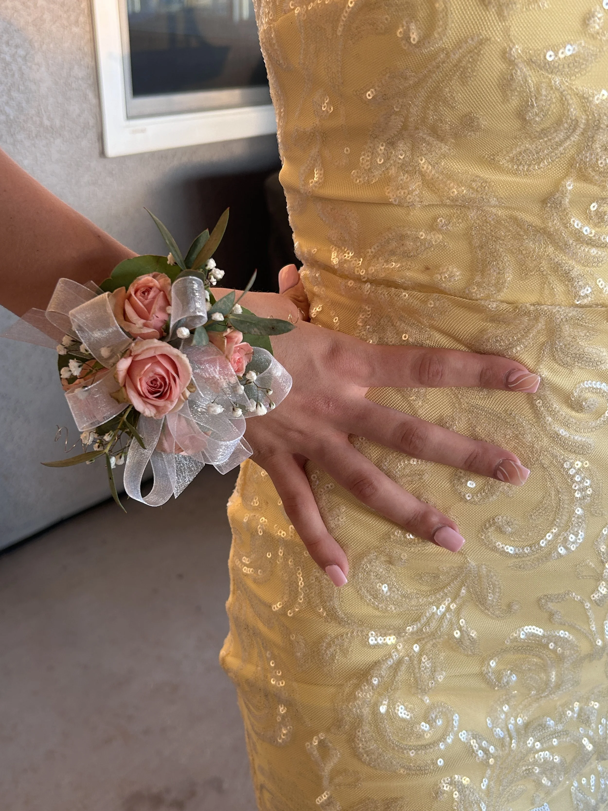 A person's hand resting on their yellow, sequined dress with swirl patterns, wearing a pink rose corsage with white ribbon and small white flowers.
