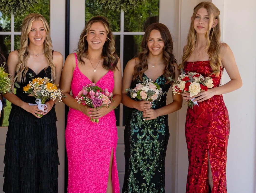 Four young women in formal dresses holding bouquets of flowers, standing indoors near a window with green foliage outside.