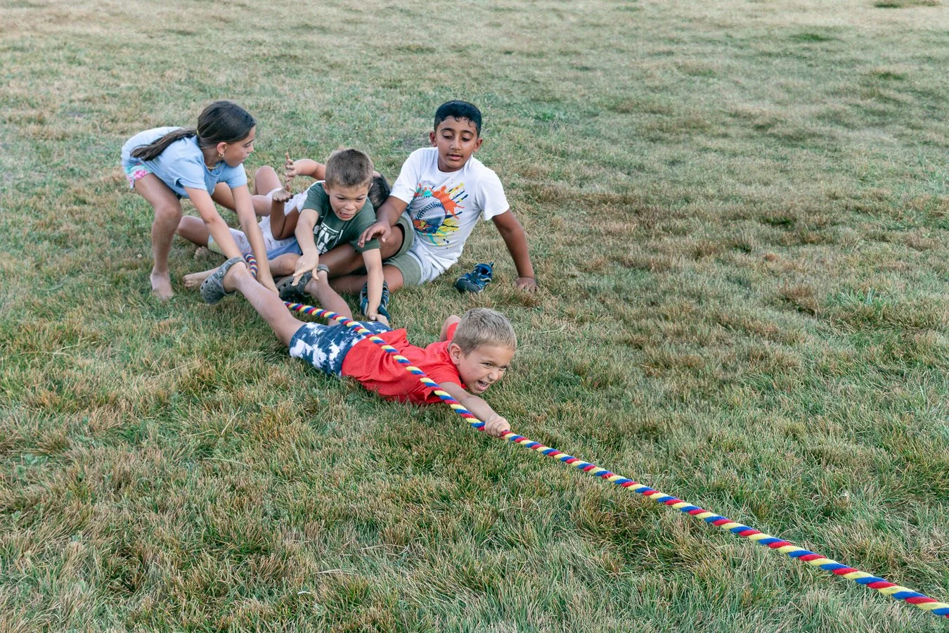 Five children playing tug of war on a grassy field, with one boy in red shirt and shorts pulling on the rope while others push and pull from the opposite side.