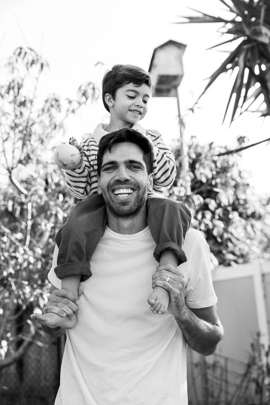 A happy man carrying a young boy on his shoulders outdoors in a garden, smiling at the camera, with trees and a birdhouse in the background.