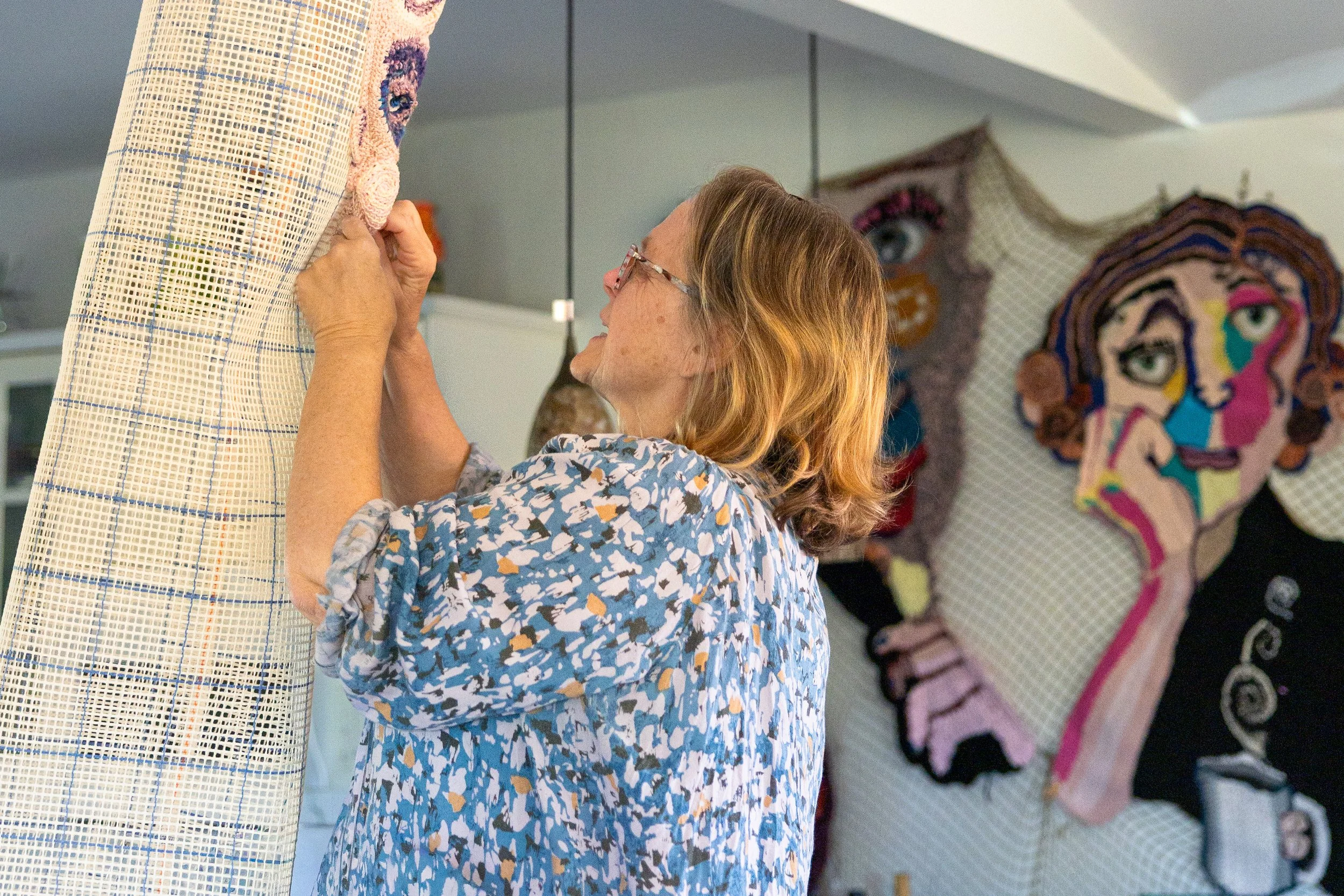An elderly woman with blonde hair and glasses is working on a craft project, attaching a piece of fabric with embroidery or appliqué details onto a vertical surface. In the background, there are colorful textile art pieces depicting stylized faces ma