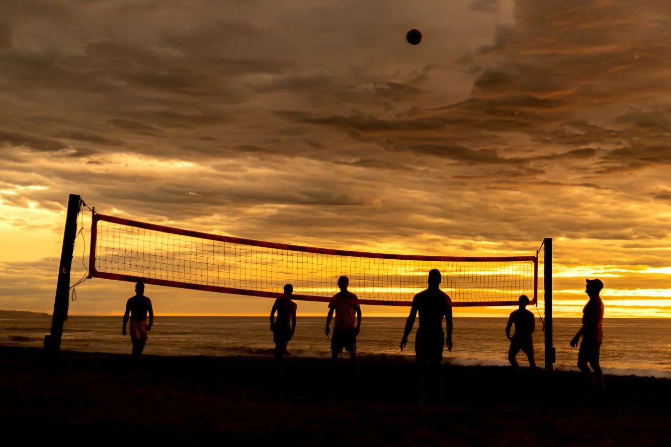 Silhouettes of six people playing volleyball on a beach at sunset with a cloudy sky and a dark moon in the sky.