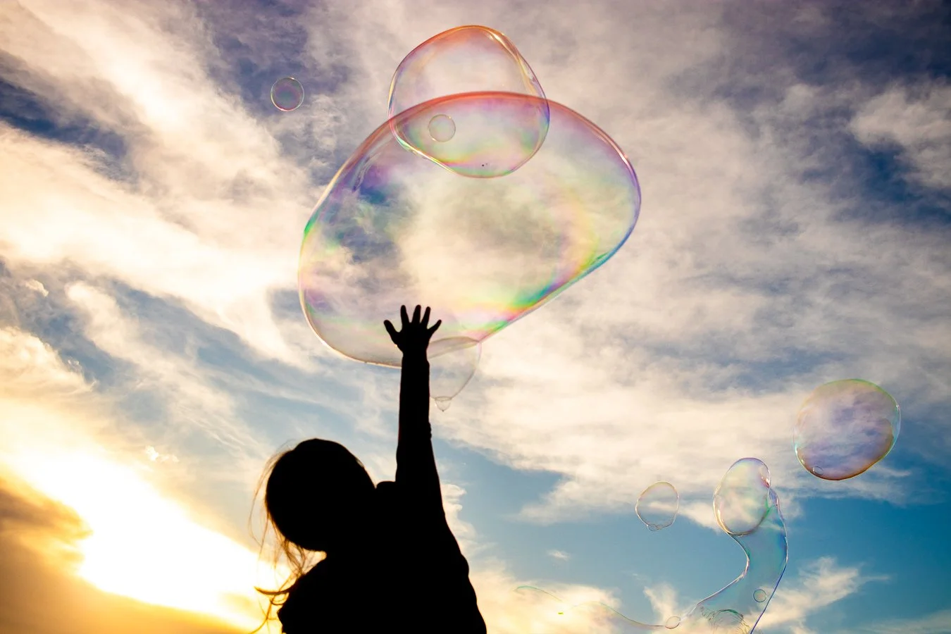 A silhouette of a girl reaching up towards iridescent soap bubbles floating in the sky during sunset.